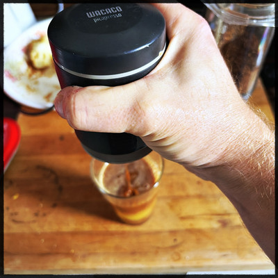 A person holding a black cylinder, which is a Wacaco PicoPresso espresso maker. That's pouring espresso into a glass of orange juice, which sits on a wooden board on a tabletop. 