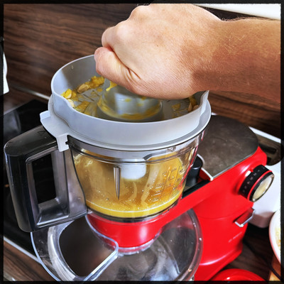 A person using a juice squeezer on a red kitchen machine. The container is already filled with orange juice a little bit. 