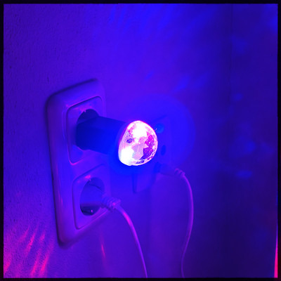 A small USB Power supply sitting in a power socket. I tiny disco light is plugged into the USB port and shining blue and purple  light. Projecting onto the surrounding wall. 