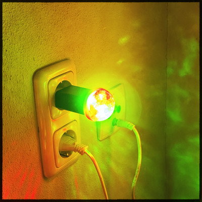 A small USB Power supply sitting in a power socket. I tiny disco light is plugged into the USB port and shining bright red, yellow and green light. Projecting onto the surrounding wall. 