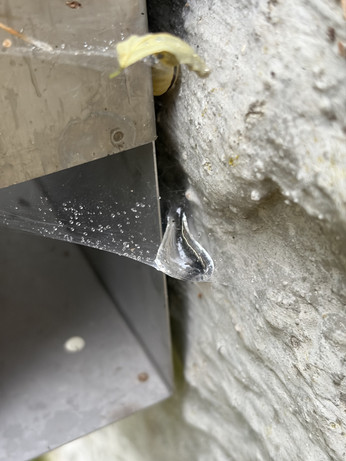 A tiny spiderweb on a metal box mounted on a concrete wall and there's a large drop of water suspended in the spiderweb